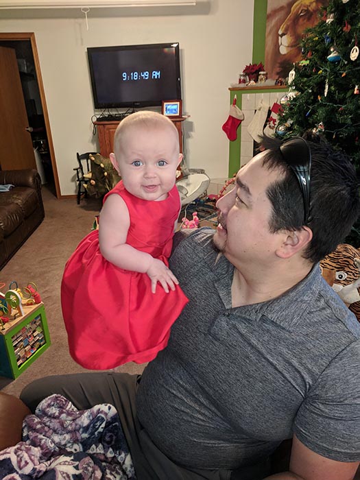 Baby Ella in her Christmas dress in the living room Dad is looking at her and holding her up.