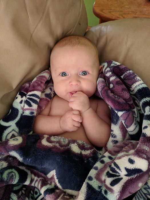 Baby Ella looking at the camera and smiling.  She is wrapped in a purple, white and blue blanket.