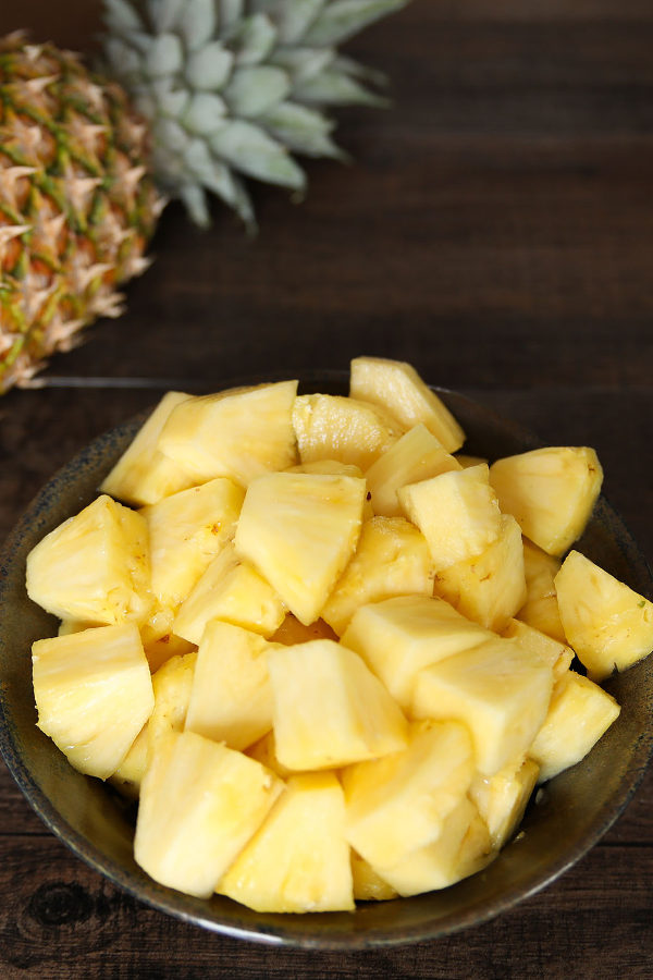 This picture shows one of the three ways to cut a pineapple in this tutorial. A pineapple sits next to a pottery bowl of pineapple chunks on a brown table.
