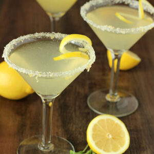 Three stunning lemon drop martinis with twists and sugared rooms. The table is decorated with a few lemons.