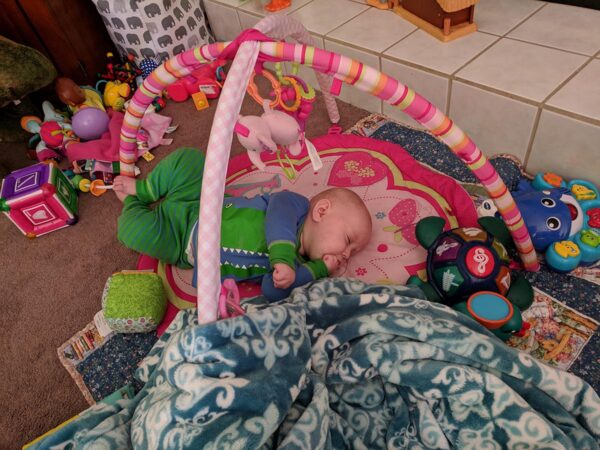 Baby Ella asleep on her play mat in Alligator pajamas.  She is surrounded by toys.