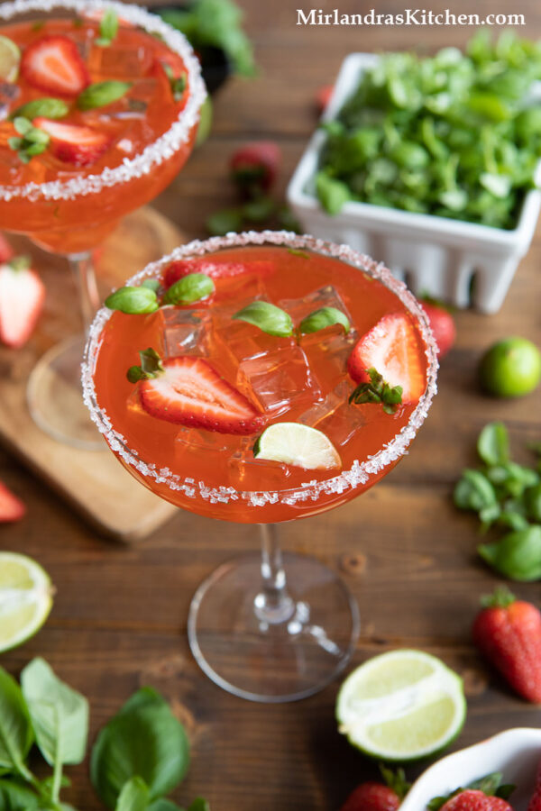 A large strawberry basil margarita is in the foreground. Around the drink are ingredients such as fresh limes and strawberries. 