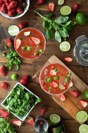 Two beautiful strawberry basil margaritas on the rocks sit surrounded by berries, limes, and basil microgreens.