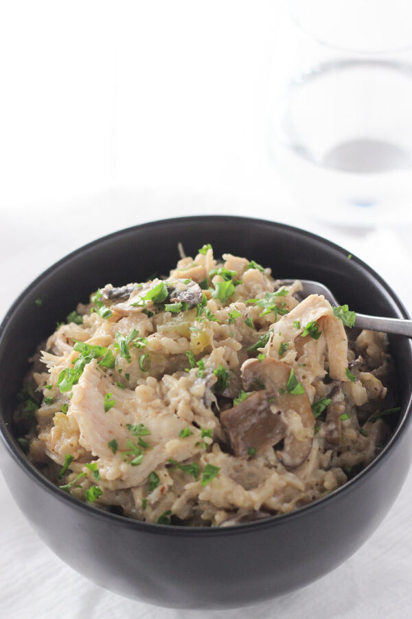 A black bowl is full if instant pot chicken and rice. You can see creamy rice, large chunks of shredded chicken, slices of mushrooms and even some celery. The dinner is garnished with fresh parsley.