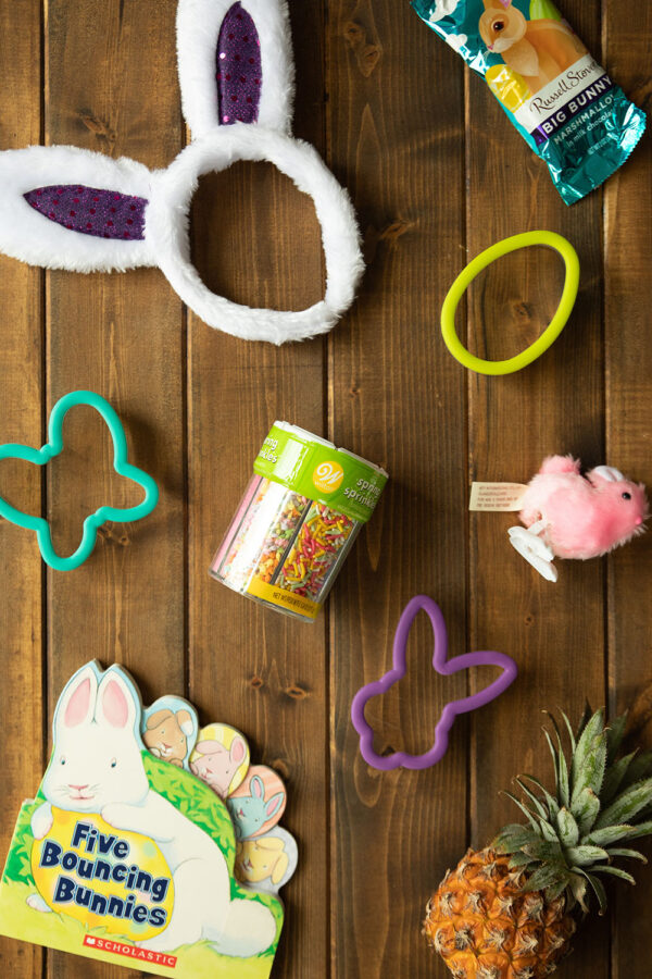 Bunny ears, chocolate, cookie cutters, sprinkles and small gifts are set out on a wooden table ready to go into a toddler Easter basket.