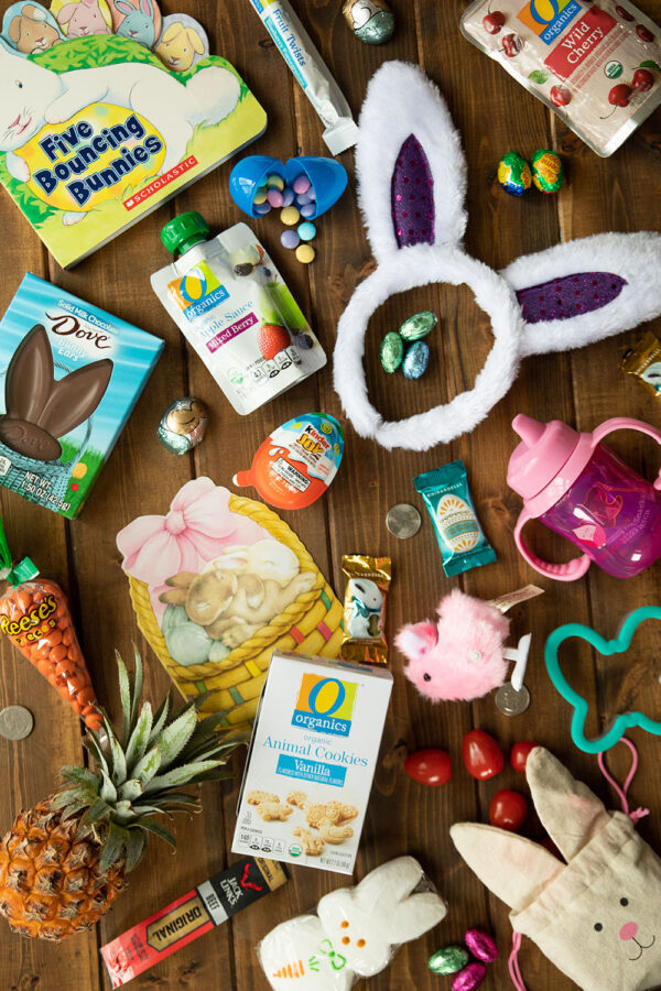 Bunny ears, a board book, and various edible treats are arranged on a wooden table ready to fill an Easter basket for a younger toddler.
