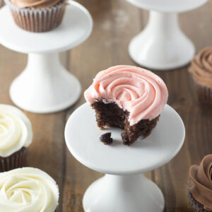Chocolate cupcakes sit on white pedestals and are scattered around on wooden table. Some are frosted in chocolate buttercream, some in vanilla and some in strawberry buttercream.