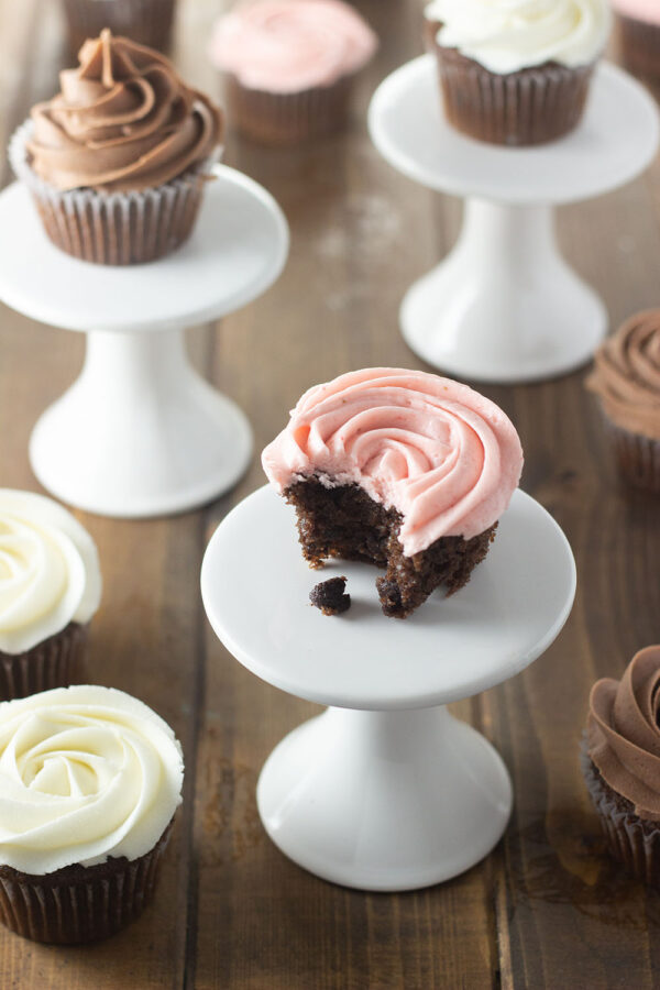 Chocolate cupcakes sit on white pedestals and are scattered around on wooden table. Some are frosted in chocolate buttercream, some in vanilla and some in strawberry buttercream.