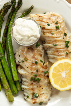 A filet of grilled cod is on a white plate next to grilled asparagus and half a fresh lemon.