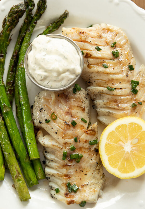 Two fillets of grilled cod sit on a white plate. There is some grilled asparagus on the plate, some tartar sauce, and a half a lemon.