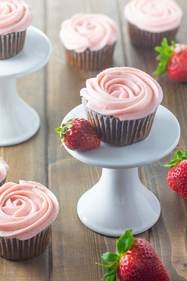 Chocolate cupcakes are frosted in fresh strawberry buttercream. One cupcake sits on a single cake stand. Additional cupcakes and strawberries are scattered around.