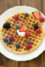 A round buttermilk waffle sits on a white plate. There are blackberries, blueberries, raspberries and strawberries on the waffle along with some whipped cream and powdered sugar.