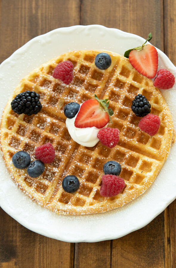 A round buttermilk waffle sits on a white plate. There are blackberries, blueberries, raspberries and strawberries on the waffle along with some whipped cream and powdered sugar.