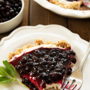A slice of huckleberry cheesecake is plated on a white plate with a fork and a sprig of mint. You can see the rest of the cheesecake and some huckleberries in the background.