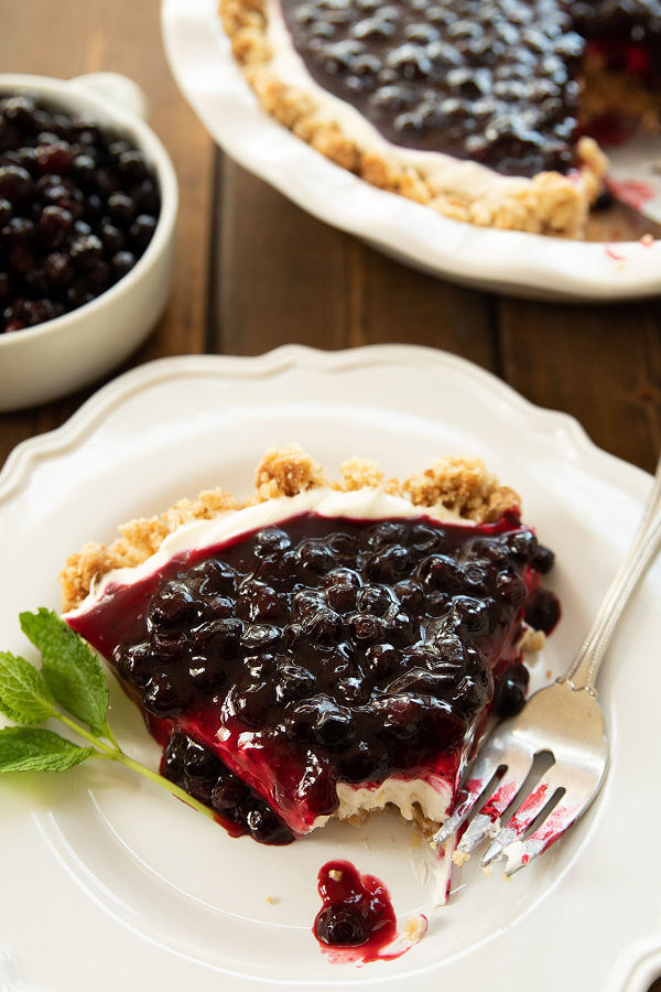 A slice of huckleberry cheesecake is plated on a white plate with a fork and a sprig of mint. You can see the rest of the cheesecake and some huckleberries in the background.