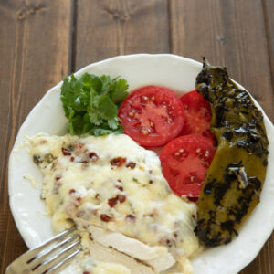 A plate of dinner features a big serving of hatch chile chicken bake, some sliced tomatoes and a whole roasted hatch chile.