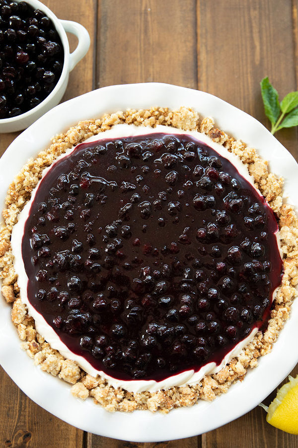 A round huckleberry cheesecake is in a white pie plate. You can see a thick layer of fresh huckleberry topping on top of the cheesecake.