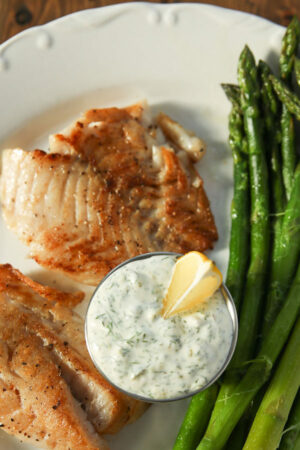 A big white dinner plate is full of tartar sauce, fried fish, and spears of asparagus. There is a wedge of lemon in the tartar sauce.