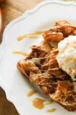 A big square of bread pudding on a white plate. It has a scoop of vanilla ice cream on top and caramel sauce drizzled over. There are apples in the background.