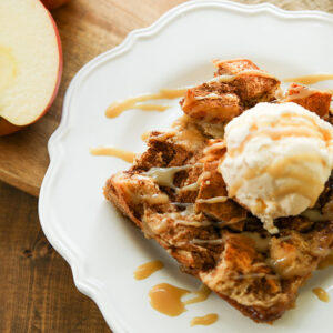 A big square of bread pudding on a white plate. It has a scoop of vanilla ice cream on top and caramel sauce drizzled over. There are apples in the background.