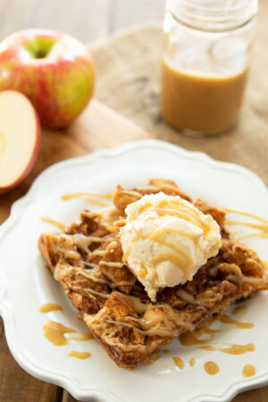 A big square of bread pudding on a white plate. It has a scoop of vanilla ice cream on top and caramel sauce drizzled over. There are apples in the background.