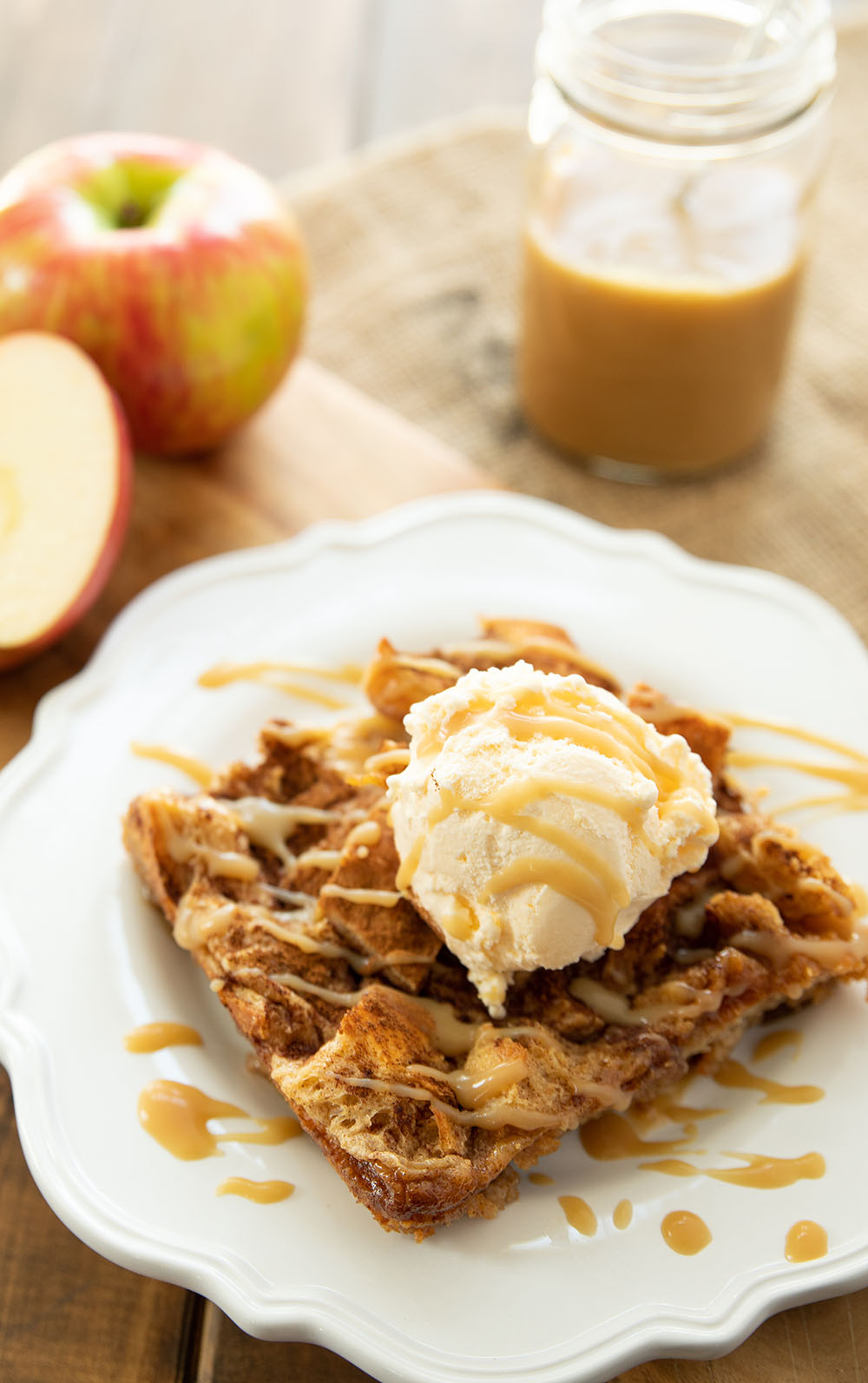 A big square of bread pudding on a white plate. It has a scoop of vanilla ice cream on top and caramel sauce drizzled over. There are apples in the background.