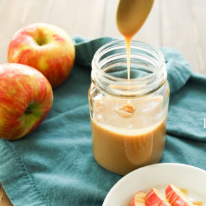 A mason jar full of homemade caramel sauce sits on a blue cloth napkin. Apples drizzled in caramel sauce sit on a white plate by the jar.