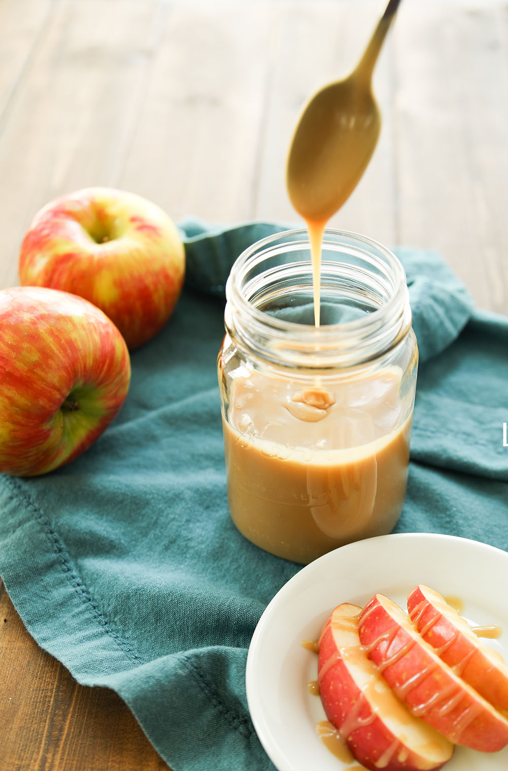 A mason jar full of homemade caramel sauce sits on a blue cloth napkin. Apples drizzled in caramel sauce sit on a white plate by the jar.