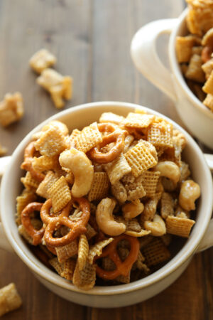 Two white bowls are full of caramel chex mix. You can see pretzels and cashews in the mixture!