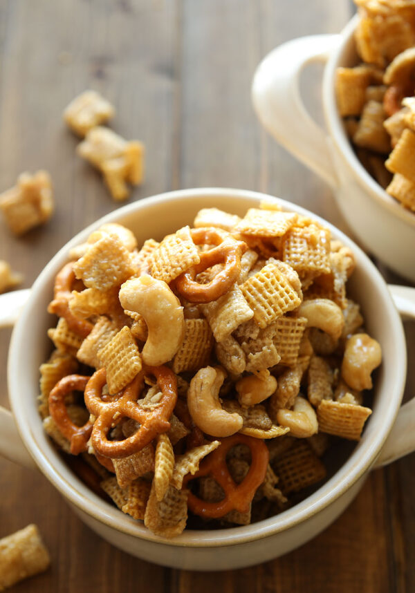 Two white bowls are full of caramel chex mix. You can see pretzels and cashews in the mixture!