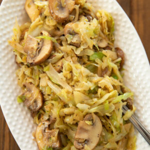 A white oval platter is full of shredded, fried cabbage with large slices of sauteed mushrooms.