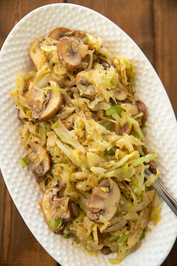 A white oval platter is full of shredded, fried cabbage with large slices of sauteed mushrooms.