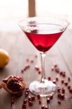 A single pomegranate martini sits on a wooden table surrounded by pomegranate seeds.