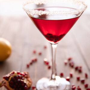 A single pomegranate martini sits on a wooden table surrounded by pomegranate seeds.