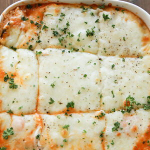 A square white baking dish is full of hot, keto zucchini lasagna. There is lots of mozzarella cheese and a little parsley on top.