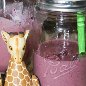 A mason jar sits on a counter next to a wooden giraffe. The jar is half through of purple, berry smoothie with a green straw in it.