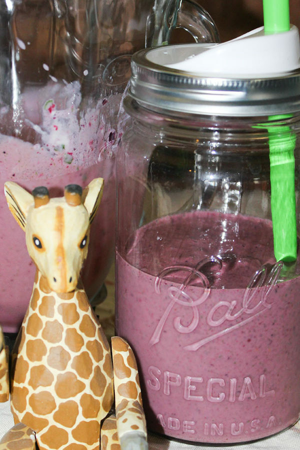 A mason jar sits on a counter next to a wooden giraffe. The jar is half through of purple, berry smoothie with a green straw in it.