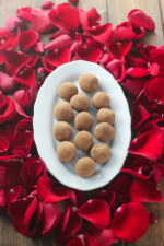 A wooden table is covered with red rose petals. In the center is a plate of hand made chocolate truffles.