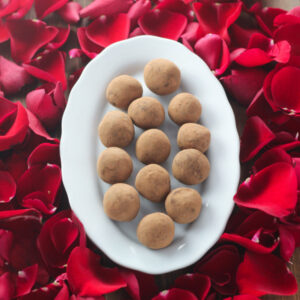 A wooden table is covered with red rose petals. In the center is a plate of hand made chocolate truffles.