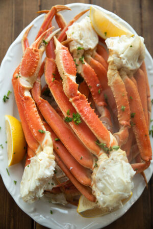 A large white platter is stacked high with steamed snow crab legs. A dusting of parsley and some wedges of lemon garnish the dish.