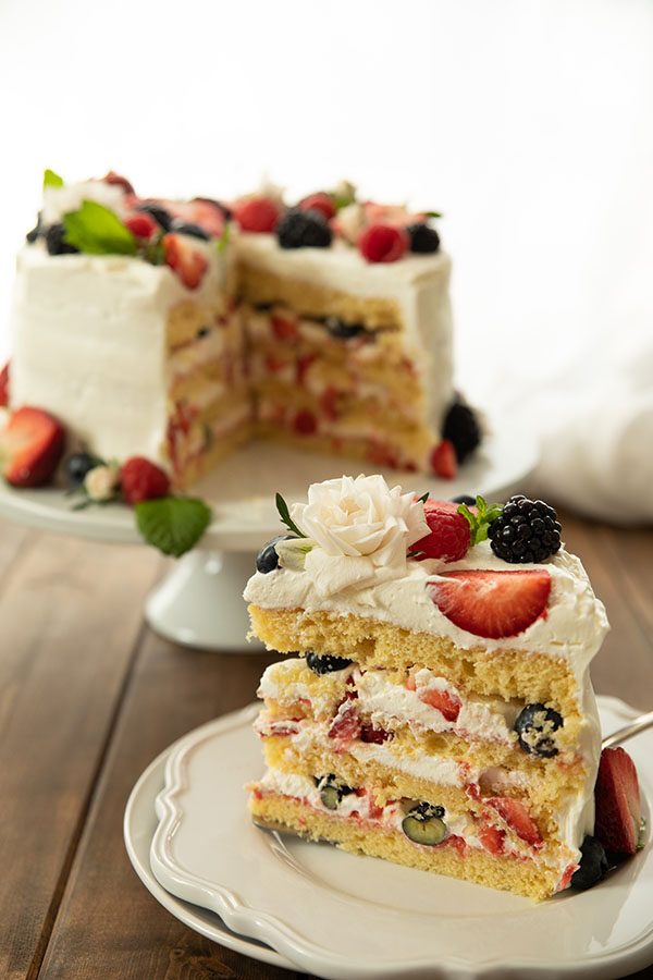 A big wedge of easy berry Chantilly cake on a white plate.  The cake is decorated with a white spray rose and fresh berries.  You can see the beautiful layers of cake and cream and berries in this photo.