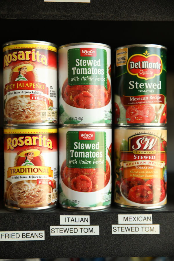 Image of pantry staples on shelf. There are cans of tomatoes and cans of refried beans.