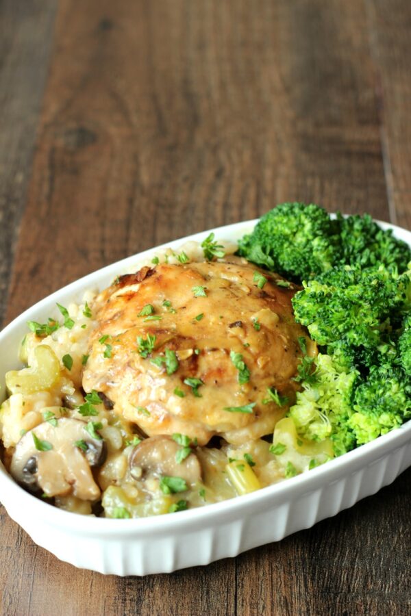 A dish of chicken and rise casserole sits on a table. The chicken is golden brown on a bed of rice with mushrooms.  There is a side of broccoli included.