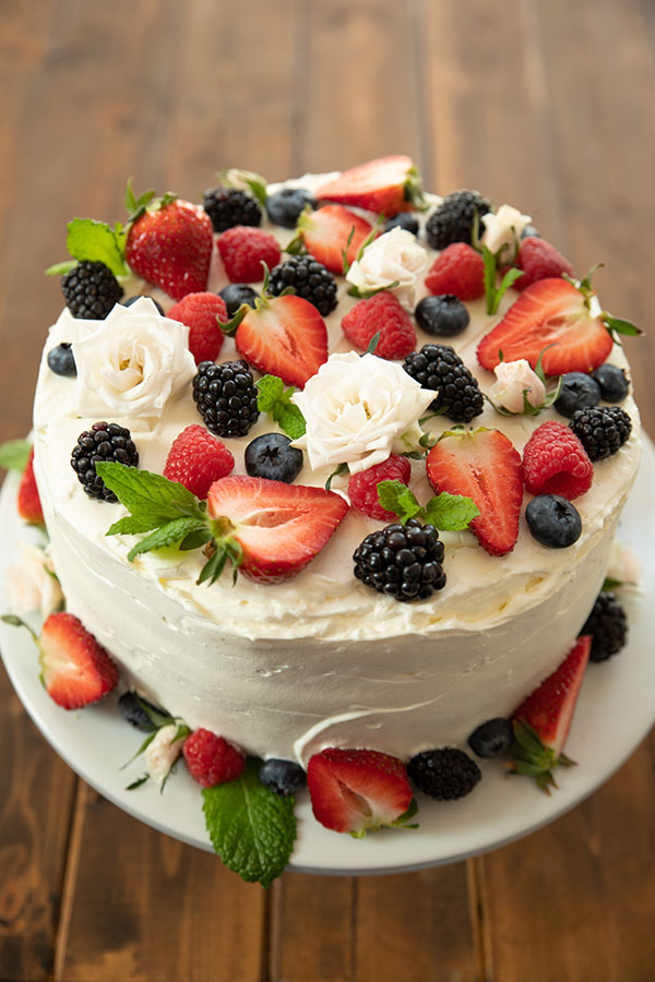 A vibrant berry chantilly cake sits on a white cake pedestal on a wooden table. The cake is covered with fresh berries and white spray roses.