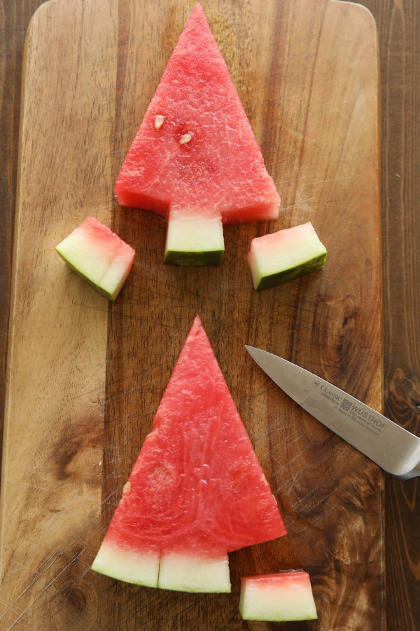 Two triangles of watermelon sliced on a cutting board.  Each shows pieces of the rind removed to show the process of cutting triangles of watermelon into trees. 