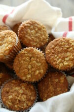 A basket is lined with a white cloth with red striped edges. It is full of golden oatmeal muffins.