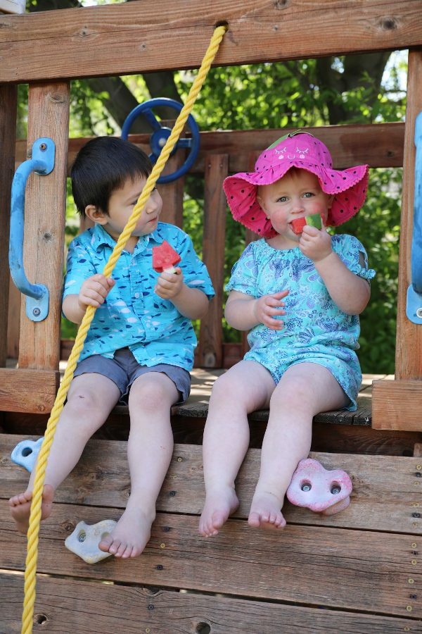 Jack and Ella sitting in their fort eating melon. Ella is wearing a strawberry hat.