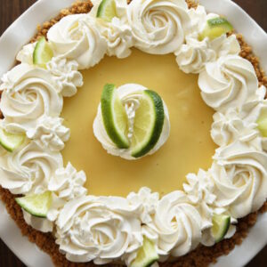 an authentic key lime pie in graham cracker crust. The whipped cream is swirled on and wedges of lime garnish the pie.