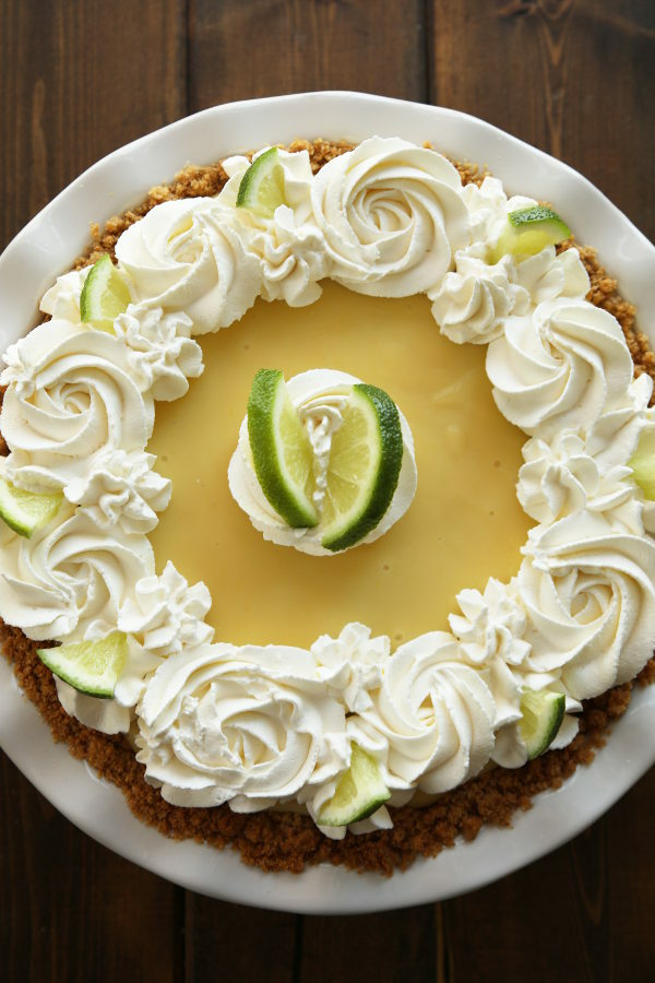 This easy key lime pie is in a white pie plate. You can see the graham cracker crust peaking out at the edges. Swirls of whipped cream and slices of fresh lime top the pie.