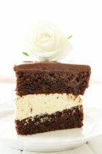 A large decadent slice of chocolate cake sits on a white plate. The cake is slathered with chocolate ganache and filled with vanilla mousse filling. There is a white rose on top of the cake.